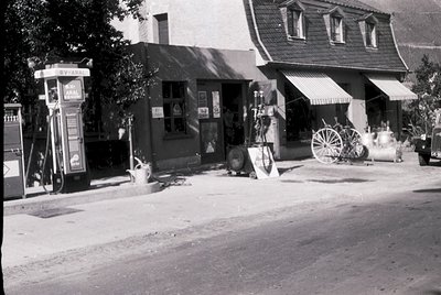 Mid-20th century American roadside scene: vintage **Arco** gas pumps beside a small **general store** with awnings, wooden wa...