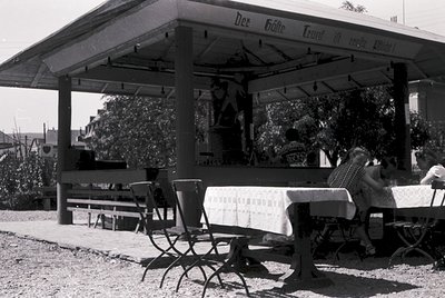 Mid-century outdoor café pavilion with sign reading *"Der Gäste Trunk in fröhlicher Gesellschaft"* (German for "Guests drink ...