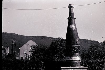 Vintage wine bottle monument with "Keller Hauser" label, mounted on a stone pedestal in a rural setting. Surrounding building...
