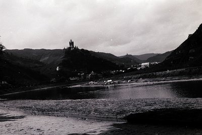 Black-and-white riverside scene featuring a medieval castle perched atop a hill, likely **Burg Eltz** (Rhineland-Palatinate, ...