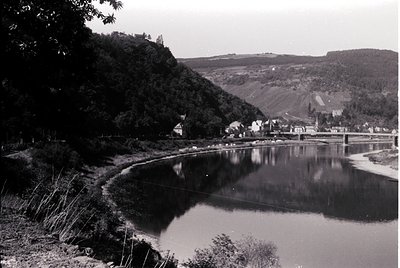 Black-and-white landscape featuring a serene river winding through a rural valley. Gentle curves mirror buildings and foreste...