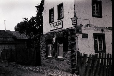 Mid-20th century German village inn with stone façade and wooden sign reading "Gastwirtschaft." Cobblestone street and rustic...