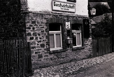 Stone-framed 1960s-70s German *Gastwirtschaft* ("Toni Schneider") with rustic cobblestone street. Black-and-white shot highli...