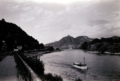 Mid-20th century black-and-white river scene with a ferryboat crossing, flanked by lush greenery and a distant hilltop castle...