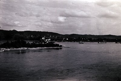 Black-and-white coastal scene featuring a calm body of water with a rocky shoreline and sparse vegetation. Distant buildings ...