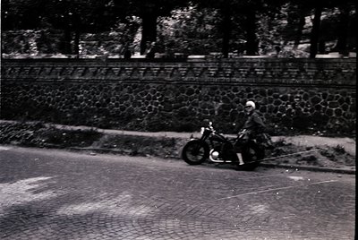 Vintage black-and-white photo of a man riding a classic motorcycle on a cobblestone road, flanked by a stone wall and dense f...
