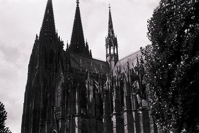 Gothic cathedral façade featuring three towering spires with intricate tracery and vertical buttresses. Black-and-white monoc...