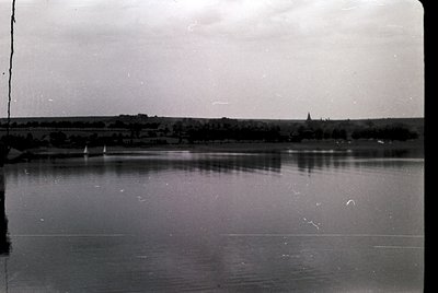 Vintage black-and-white photo of a serene lakeside village with low-lying buildings and a prominent church spire. Calm water ...