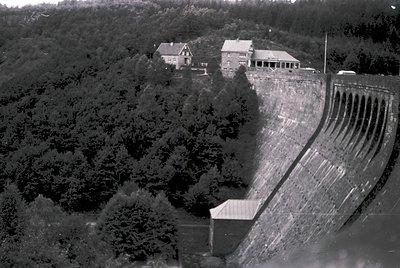 Aerial view of a mid-20th century concrete dam with adjacent buildings perched on forested hills. Architectural style suggest...