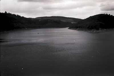 Black-and-white landscape of a serene lake bordered by forested hills, captured in mid-20th century style. Minimal human pres...