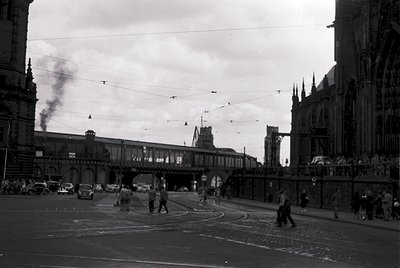 Black-and-white street scene featuring a historic European railway station with Gothic Revival architecture, visible smoke fr...