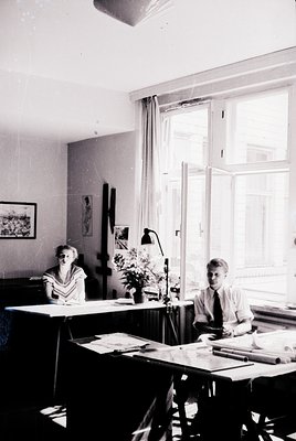 Mid-20th century office interior with two professionals seated at desks, likely 1950s–1960s. Woman in striped blouse reviews ...