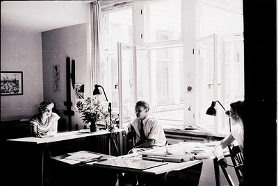 Mid-century study with three individuals engrossed in work, framed by large windows and natural light. Wooden desks, vintage ...