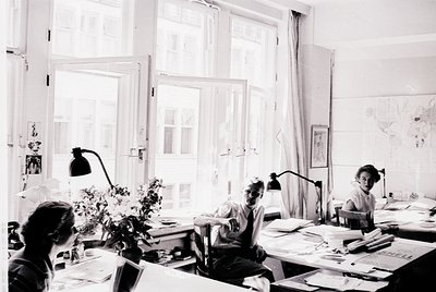 Brightly lit 1950s–60s office with three focused individuals at desks. Natural light floods through large windows; vintage de...