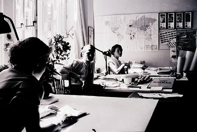 Three women collaborate in a mid-century office, surrounded by maps, fabric swatches, and rolled textiles. Natural light floo...