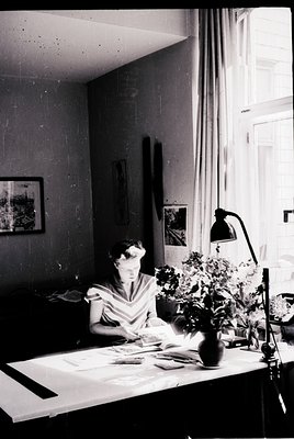 Mid-century study scene featuring a woman engrossed in drafting or sewing, seated at a wooden desk under natural light. Decor...