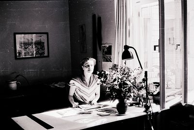 Mid-century woman seated at a desk in a sunlit study, writing or drafting. Vintage desk lamp, framed artwork, and floral arra...