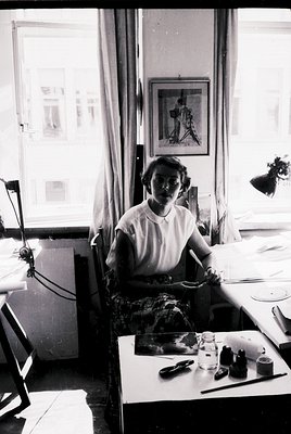 Vintage black-and-white portrait of a woman seated at a drafting table in a sunlit studio, likely mid-20th century. She wears...