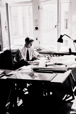 Mid-century office scene featuring a man in a suit and tie engrossed in drafting at a large drafting table. Surrounding mater...