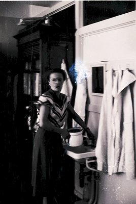 Vintage black-and-white kitchen scene featuring a woman in a striped apron holding a mug, standing beside a vintage stove wit...