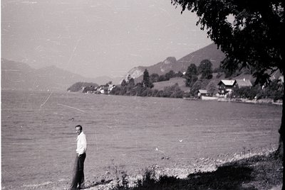 Vintage black-and-white lakeside scene featuring a man in mid-20th-century attire (vest, tie, trousers) standing by calm wate...