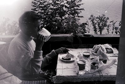 A man in mid-20th-century attire drinks from a cup while seated at a table laden with small ceramic dishes, jars, and a cover...