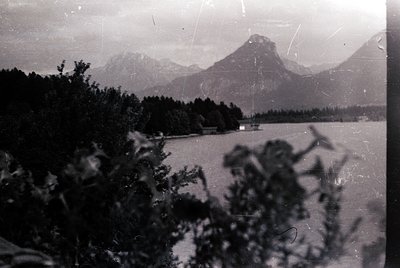 Vintage sepia-toned alpine lake scene with rugged peaks in background. Dense forest framing left edge suggests early 20th-cen...