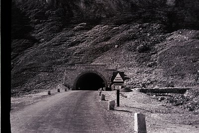 Mid-20th century tunnel entrance on a winding mountain road, marked by a 50 km/h speed limit sign. Stone tunnel arch framed b...