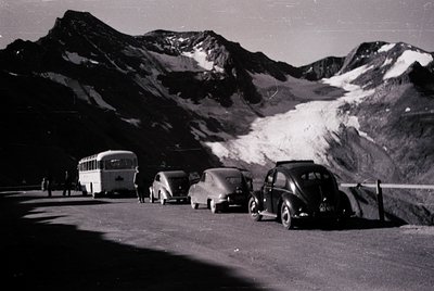 Vintage alpine road scene with vintage cars (1950s–60s) parked beside a bus on a winding mountain pass. Snow-capped peaks and...