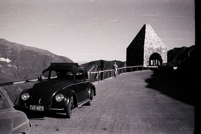 Vintage black-and-white shot of a 1950s-era Volkswagen Beetle parked beside a mountain road, with a pyramid-shaped stone stru...