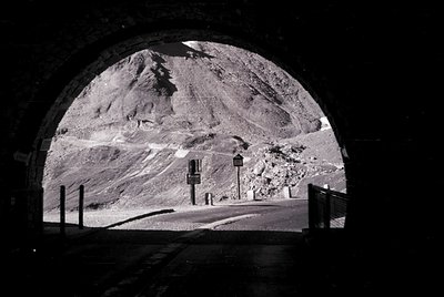 Black-and-white tunnel exit revealing rugged alpine terrain with winding road and signage. Dramatic contrast between dark tun...