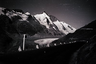 Black-and-white alpine road winding through jagged peaks, flanked by snow patches and rocky slopes. Mid-century road sign vis...