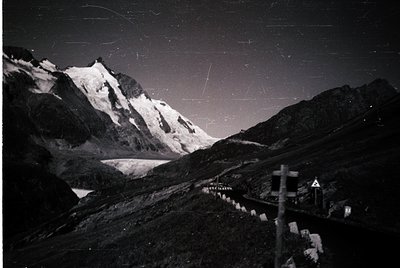 Black-and-white nighttime alpine scene with jagged peaks, illuminated by moonlight and star trails. Snow-covered slopes contr...