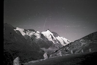 Black-and-white alpine panorama showcasing jagged peaks with snow-capped ridges under starry night sky. Long-exposure streaks...