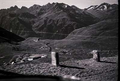 Aerial black-and-white view of rugged alpine terrain with steep, forested slopes and jagged peaks. Foreground shows a winding...