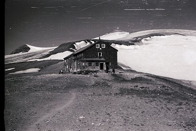 Two-story alpine chalet-style building perched on rocky terrain, surrounded by snow-capped peaks. Constructed with dark wood,...
