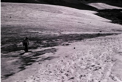 Black-and-white alpine scene showing a lone hiker on a snow-covered trail, surrounded by rugged terrain. The individual wears...