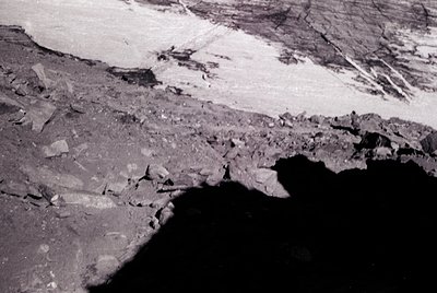 Aerial view of a glacier’s terminus meeting a dark, rocky shoreline. Contrasting textures: jagged ice and fractured bedrock. ...