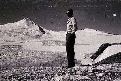 A lone individual stands atop rugged terrain, framed by expansive snow-capped peaks under a starry night sky. Mid-century out...
