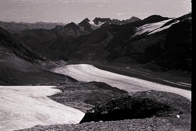 Black-and-white alpine glacier nestled between jagged peaks, likely from mid-20th century. Snow-covered valley floor contrast...