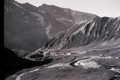Aerial black-and-white shot of a winding mountain road hugging steep slopes, flanked by rugged alpine terrain. Mid-20th centu...