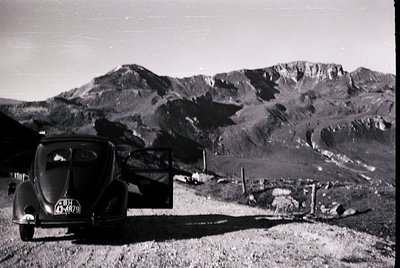 Classic 1950s-era Volkswagen Beetle parked on a rugged alpine road, framed by jagged peaks. The vintage license plate suggest...