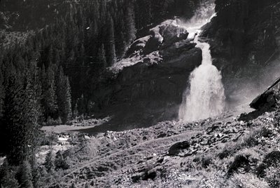 Black-and-white aerial view of a cascading waterfall plunging into a forested valley, framed by rugged cliffs. Mid-20th centu...