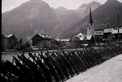 Black-and-white alpine village scene with wooden fencing and snow-covered rooftops. Prominent church steeple and sign reading...