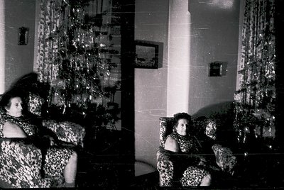 Vintage black-and-white interior shot of two women in patterned dresses seated on a sofa in a dimly lit room. Reflective glas...