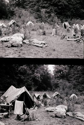 Historic black-and-white photo of a rugged campsite in a forested area, likely mid-20th century. Tents, wooden logs, and make...