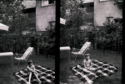 Mid-20th-century backyard scene featuring two children lounging on makeshift wooden loungers. Concrete apartment block and lu...