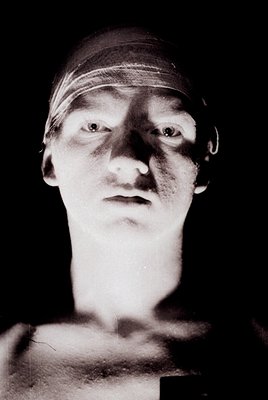 Black-and-white portrait of a person with a bandaged head, likely post-surgery or injury, wearing a white headwrap. Piercing ...