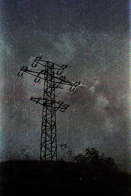 High-voltage lattice steel transmission tower with multiple cross-arms and insulators, set against a cloudy sky. Likely mid-2...