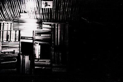 High-angle black-and-white shot of densely packed bookshelves under slatted ceiling beams, creating geometric shadows. Minima...